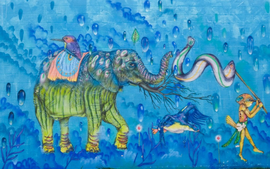 Part of a mural by Eder Muniz, on blue background, small elf like character carrying a pole with a long banner on the wnd of it, an elephant following behind, with a bird sitting on the back of the elephant looking backwards