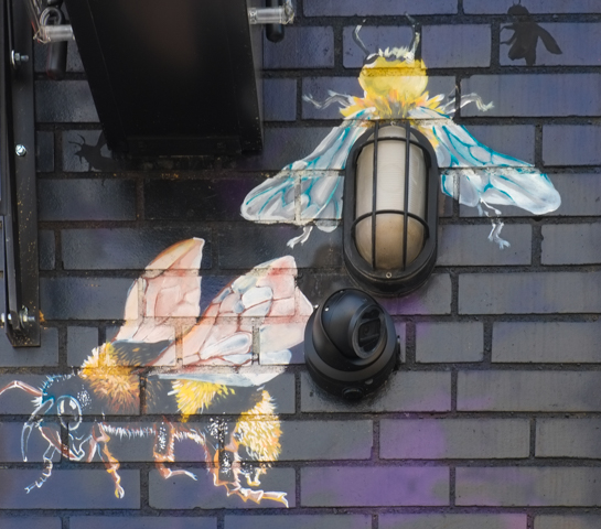part of a mural, two bees around a real light on a brick wall 