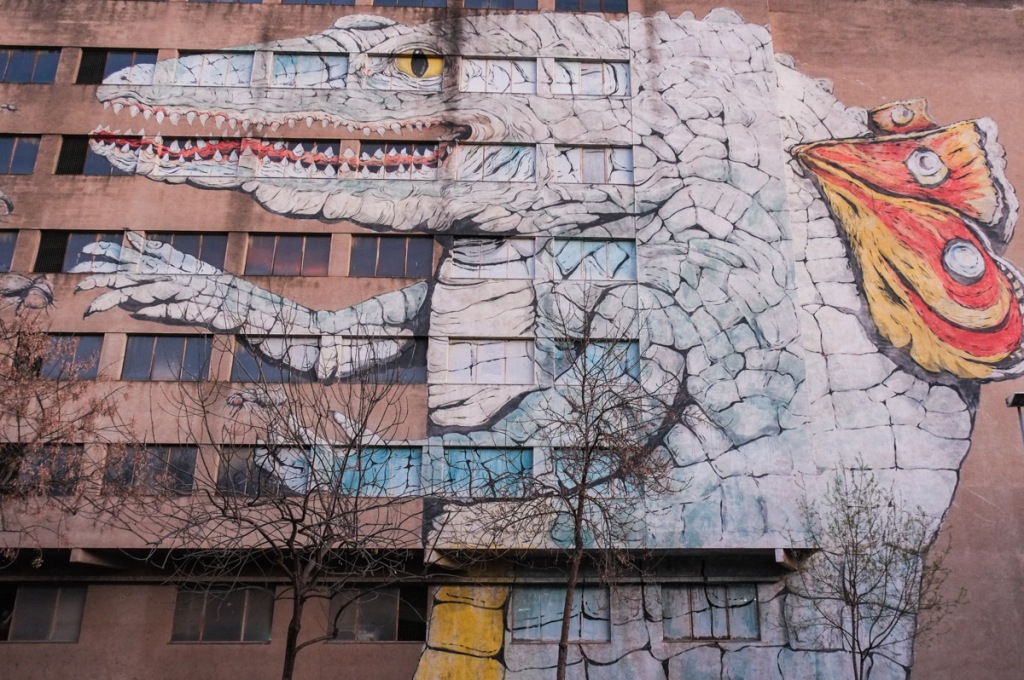 straight frontal view of ericailcane mural, mural of a large crocodile with butterfly wings on its back, standing on its hind legs, reaching for flying insects to eat, on the side of an apartment building 