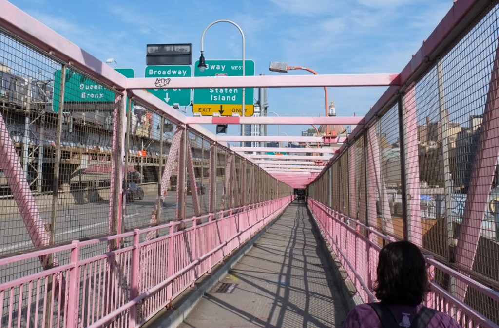 crossing the Williamsburg bridge, with pinkish red metal railings and girders, pedestrian walkway is beside and slightly above road and subway lines.