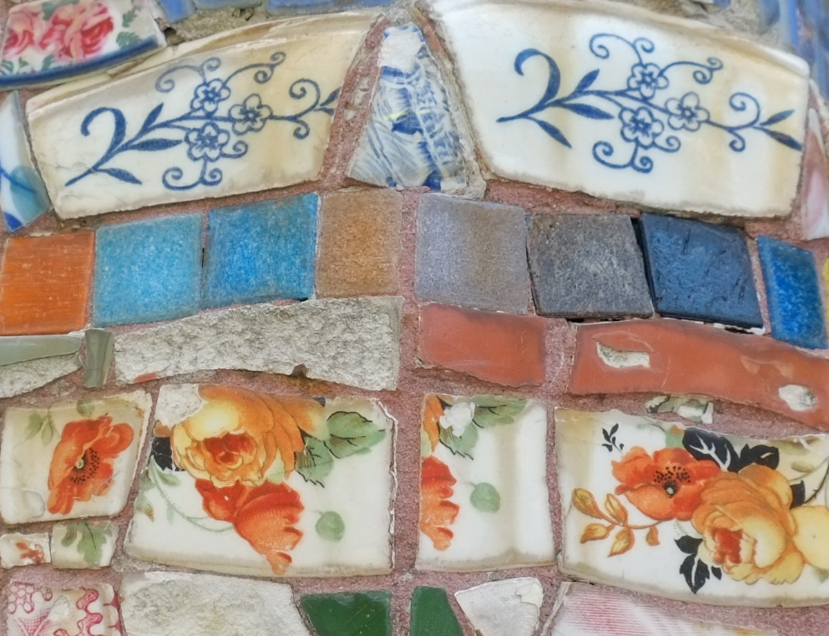 close up of a mosaic made with broken pieces of ceramic including some with pictures of blue flowers, and others with orange and red flowers, also stone like square pieces in blues and greys,