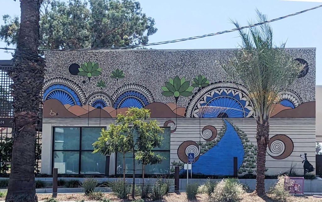 20 by 30 foot mural on the ide of a 2 storey building, with small trees in front, semi circles in blue, swirls in red, and black dots make up most of the design, incorporates two windows of the building