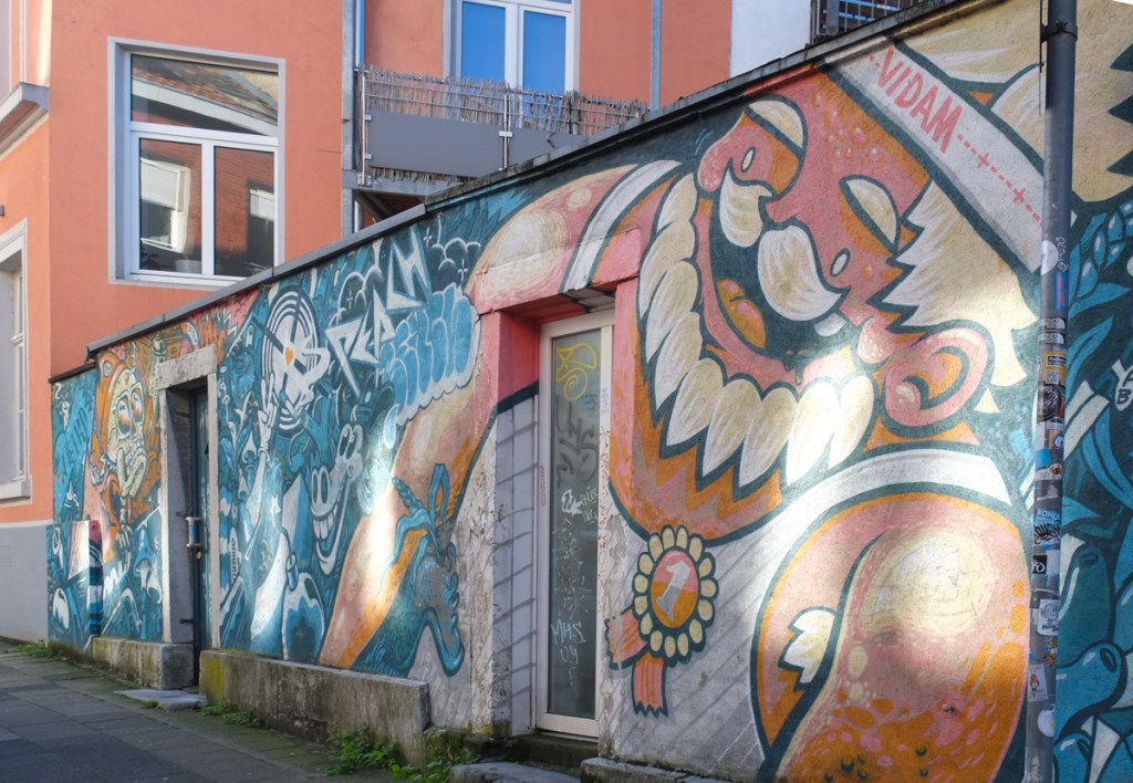 mural by vidam in aachen, large man with orange skin and white hair including mustache and beard, wearing sleeveless white top with a first prize pin on it. 