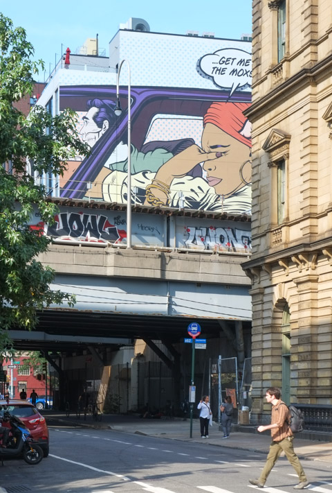 a man crosses Bedford by the bridge for the ramo to the Williamsburg bridge, a mural is above the bridge but on other side o frailway