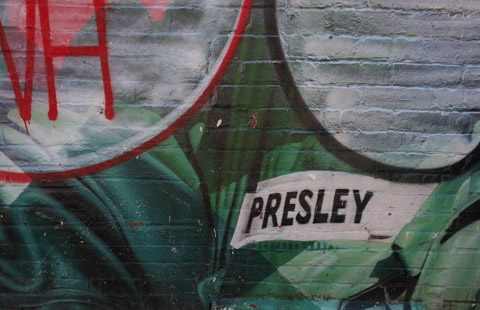 part of a defaced mural, white tag over picture of Elvis Presley, name tag on army uniform saying Presley is clearly seen