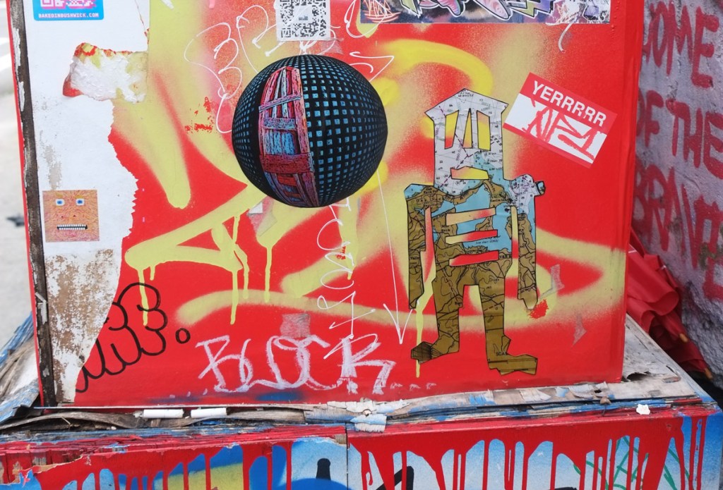 stickers and graffiti on a red metal box on a sidewalk, including a multi coloured stickman in oinkm bklue, green, and brown and a round sticker with blue square on black and a reddish stikman like shape, also a small square sticker with a face on it, also a sticker in red and white with word yerrrrr