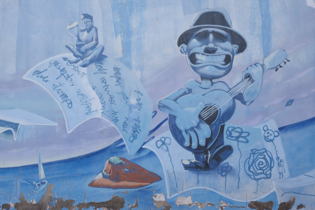 part of mural in mostly blue tones, a grinning man in a fedora plays the guitar as a sheet of paper with flower drawings on it passes by 