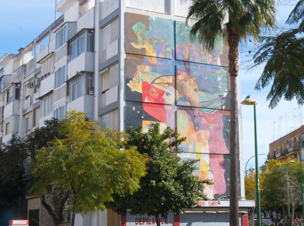 behind a few small trees, a large mural called Planeta Terra, in Seville Spain, a horse head, a pink face, and other abstract colours and shapes