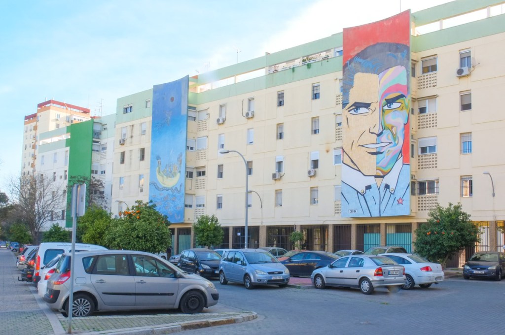 Residential buildings with murals on them, 2 large murals, and one large wall of green. One of the murals is a man's portrait