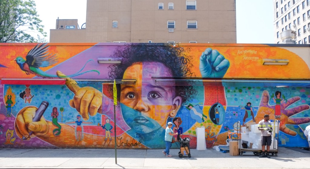 full Joel Artista and Artolution mural in East village of Manhattan, New York City, child ith short curly black hair and four arms to represent 4 activities, hand held open, holding crayon, pointing at a bird, with pedestrians walking in front of it  