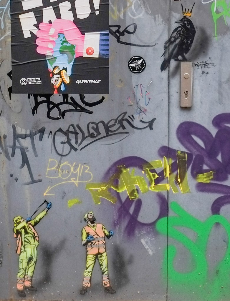 stencil graffiti of two workmen looking up at a large black bird, one of the men has a yellow sling shot that he is aiming at the bird