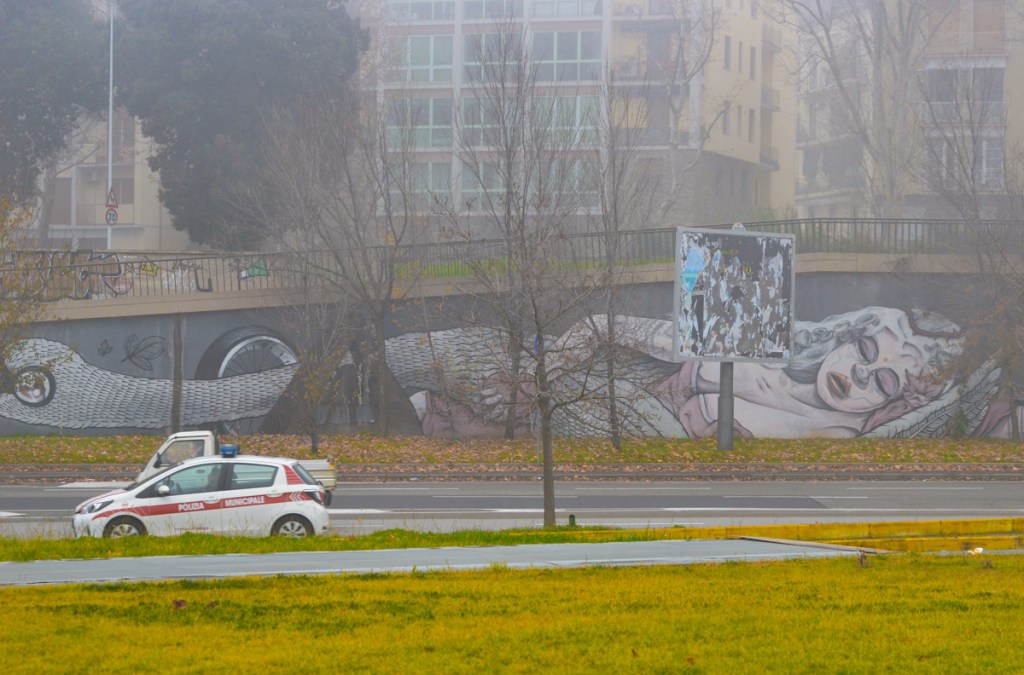 foggy day, looking across a road at a long mural on a concrete wall or ramp with railing above, mural is of a woman sleeping on the ground, head of woman on right hand side but head of swan on the left