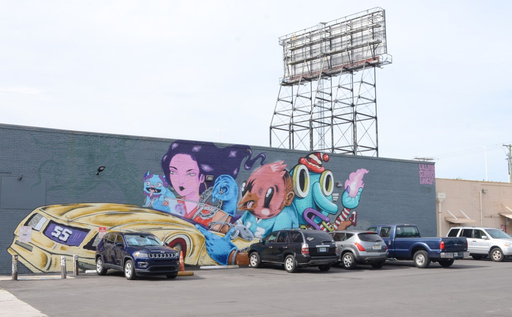 vertical mural along the side of a one storey building, grey background, a group of characters riding in a gold coloured car, painted by lillipore, sentrock and birdcap in Tampa Florida 