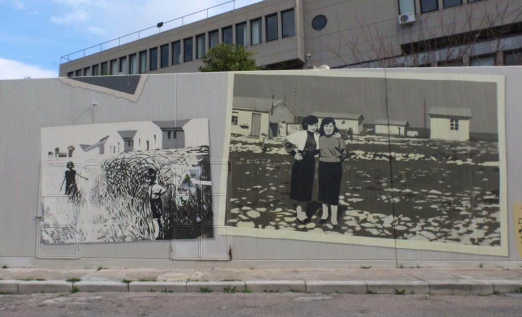 two black and white photographs of people and life at camp 65, turned into images on a mural painted on a wall, one shows two women standing in front of small white buildings 
