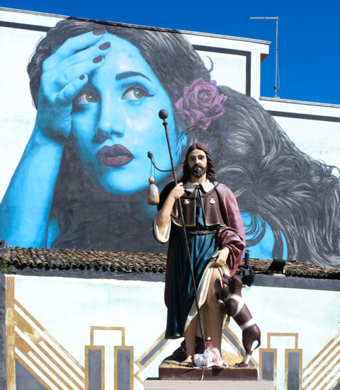 statue of a saint in a town square in Italy, with a blue mural of a woman by Leticia Mandragora behind