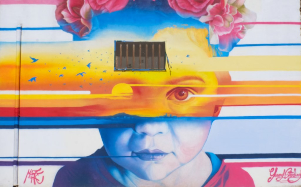mural in Stornara, young girls face, flowers in hair, yellow and blue stripes through face, one eye covered, 