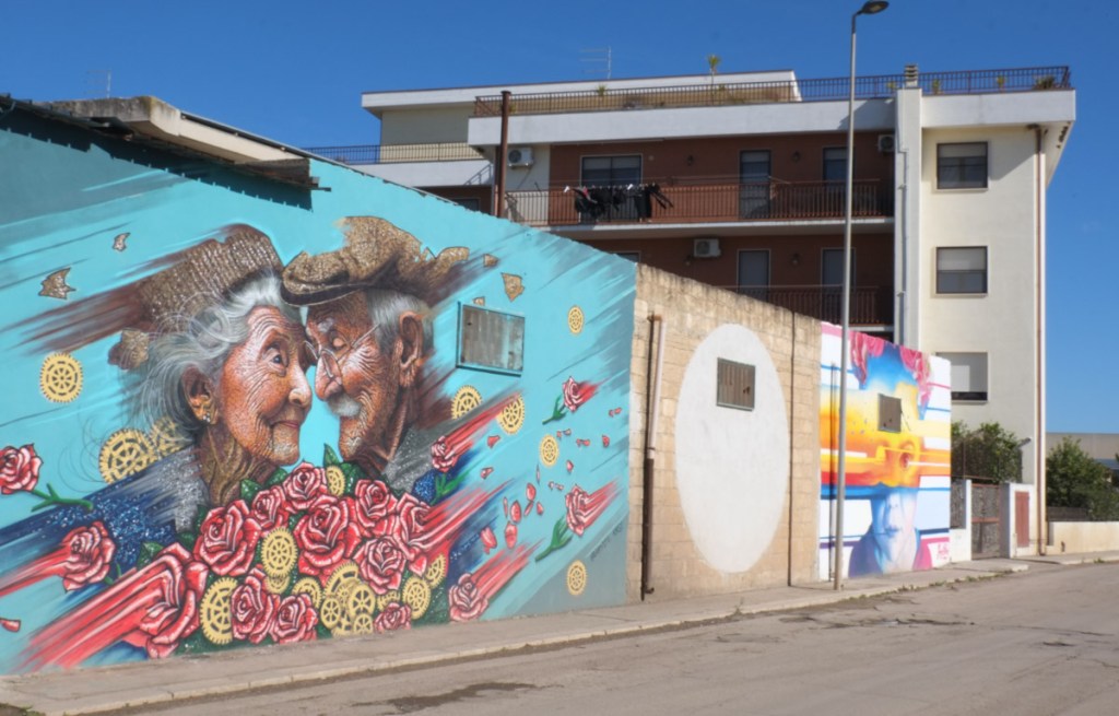 two murals in stornara, wall - older couple smiling at each other with lots of roses in the picture, called eternal love.  other mural is young child with yellow, red, and blue stripes, 