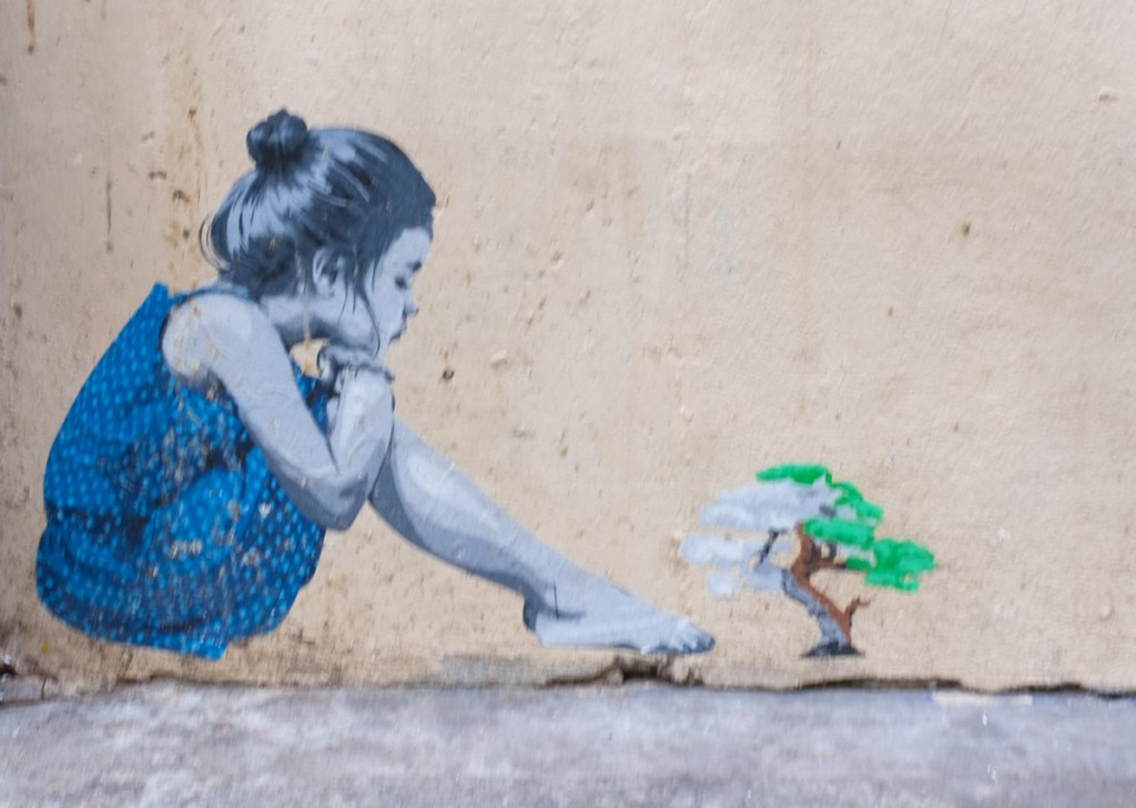 graffiti painting, girl sitting on sidewalk looking at a small tree