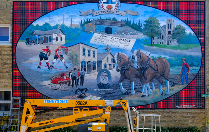 large mural, outdoors, black watchman plaid background with large interior oval containing many images of pioneer life, horses pulling a plow in a field, men playing hockey outside, a church, a wagon on a street with a couple of stores, 