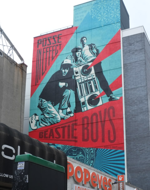 large mural by shephard fairey in the village area of new york city, featuring the music group the Beastie Boys.  An image from one of their album covers, Posse In Effect in mostly red and blue.  Men standing beside a large ghetto blaster and one man crouching beside it.  Looming over stores on 14th Ave near the corner of Avenue A. 