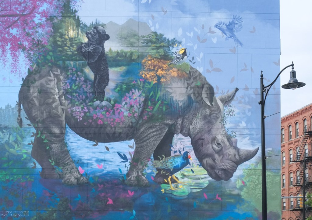 mural, blue background, a large grey rhinocerus with one horn, part of his body is a landscape scene with water, trees, flowers, and other plant life, along with a small black bear.  There are also birds.  A mural by Ernesto Maranje