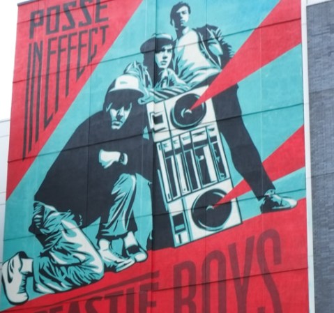 part of large mural by shephard fairey in the village area of new york city, featuring the music group the Beastie Boys.  An image from one of their album covers, Posse In Effect in mostly red and blue.  Men standing beside a large ghetto blaster and one man crouching beside it. 