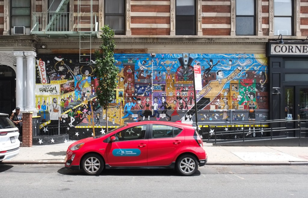 a red car is parked in front of a street art mural beside corner social store, planet harlem mural by paul deo