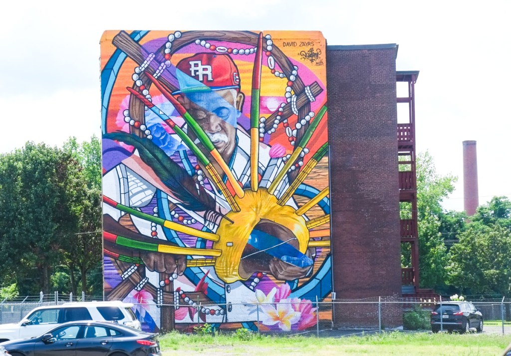 large mural on the side of a midrise residential building in holyoke, of a man in a red baseball cap who is making a mask, by david zayas and don rimx, vacant land around the building, cars parked nearby