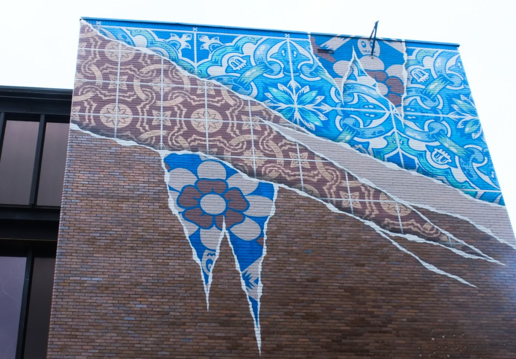 mural by add fuel, high up on two sides of a corner of a brick building, designs in blue and white as well as brown and white, painted to look like layers of wallpaper being removed to reveal the layer below,