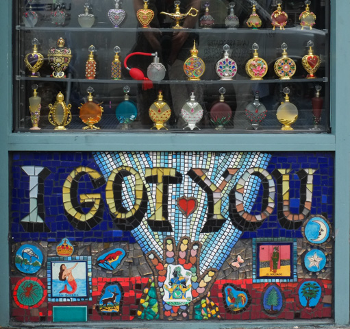 in the window of a store, shelves with display of fancy decorated perfume bottles, in gold and clear glass, under it is mosaic saying i got you, mermaid, hand, white flower with five petals, small butterfly, heart, 