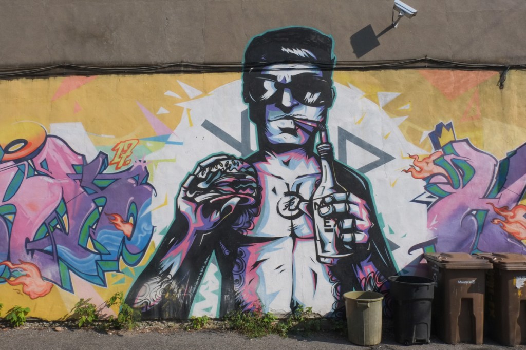 painting, street art, on side of building, black line drawing of a man with food and drink, a sandwich or burger in one hand and a bottled drink with a straw in the other