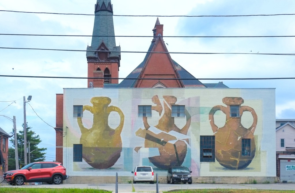 large mural by Bryan Beyung on the side of an old brick church, Elliot church, in Lowell Massachusetts. Shows three large pottery jugs, the first is whole, the middle is broken, and the last is repaired. 