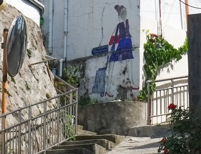 street art painting on a wall, woman in blue skirt, carrying red purse, and pulling blue shopping cart