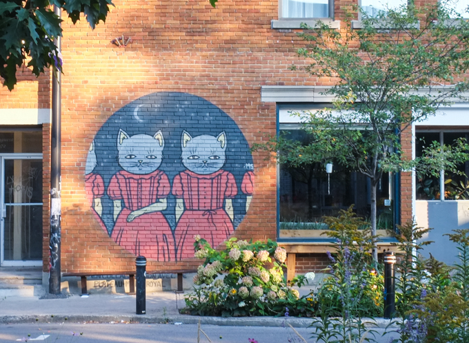 mural on a red brick wall in Montreal, circle, night with moon in the sky, two identical cats standing, wearing identical red dresses