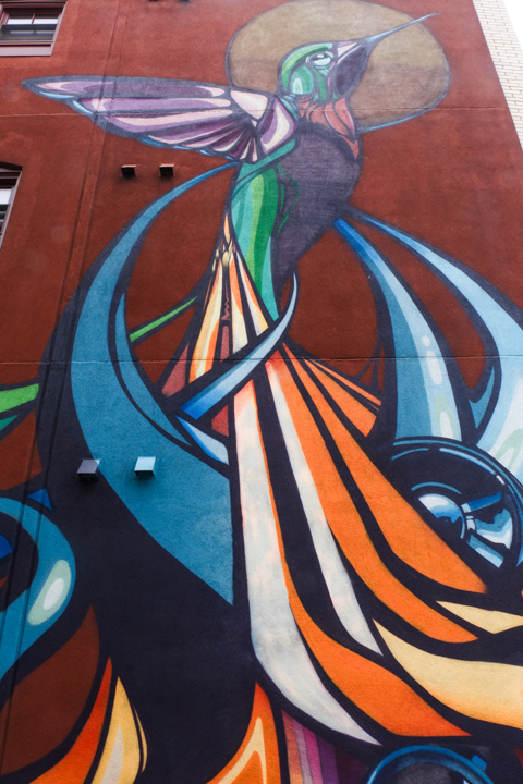 vertical mural of hummingbird, many colours, with many long flowing tail feathers