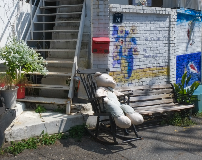 a very large stuffed teddy bear is sitting in a rocking chair on the sidewalk beside a house.  bear is weathered and faded. 