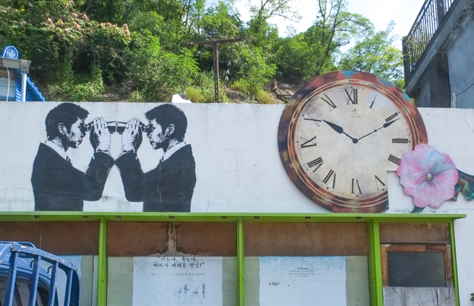 two street art paintings in black and white, on the right is an analogue clock with hands pointing to time that is 2:10.  on the left are two men with binoculars, looking at each other up closely so that the ends of the binoculars touch