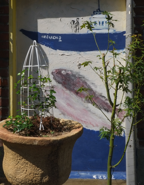 under a window and behind a planter, a rough painting of a boat with people on it, and large fish in the water below