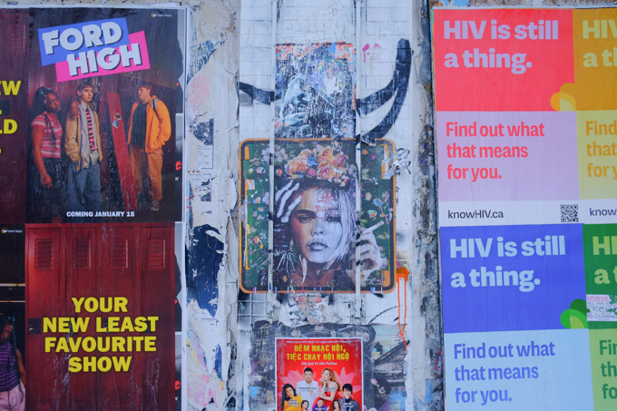posters on hoardings, some advertising television programs, some HIV education website and in the middle is a graffiti street art picture of a woman with flowers in her hair 