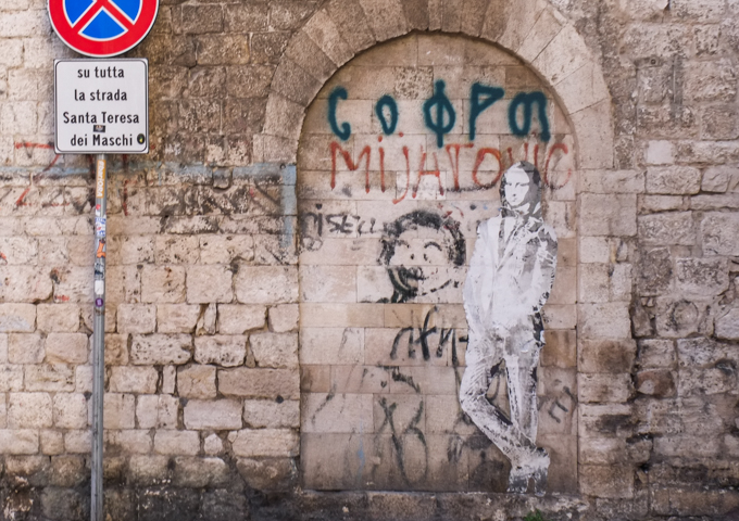 life size black and white paste up of Mona Lisa's head on a man's body wearing a suit, on a stone wall, graffiti