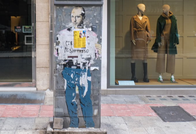 large paste up of a man in blue trousers, on a box on sidewalk, in front a womans clothing store