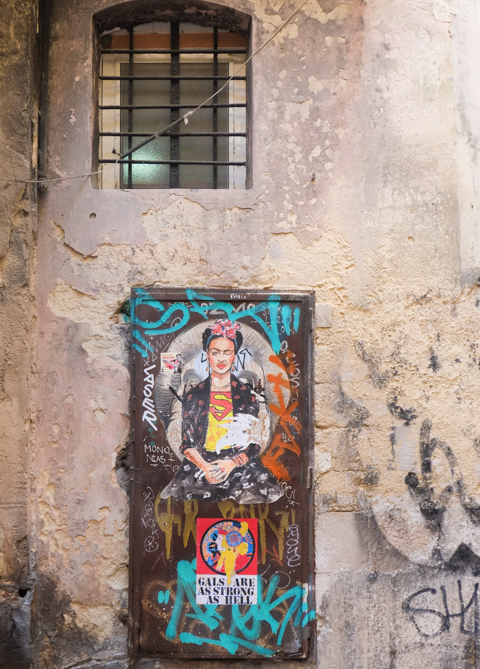 two pieces of graffiti on a wall, one is frida kahlo with superman t shirt and others is gals are strong as hell sticker