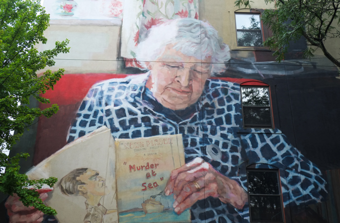 part of a mural by helen bur, an older woman sitting, holding a book called murder at sea