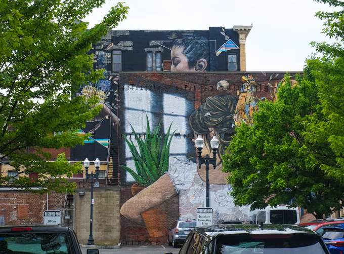 from a parking lot in Lynn, two murals on two different buildings, one partially obscured by the other. David Zayas woman in a white blouse with a rooster on her shoulder is in front