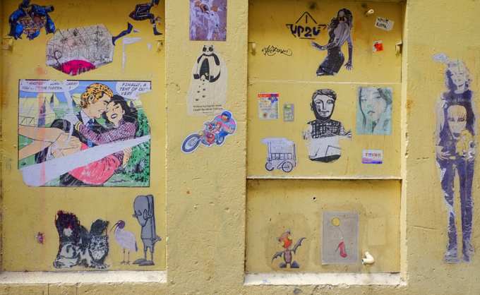 a section of yellow wall in an alley with a number of different paste up graffiti pieces