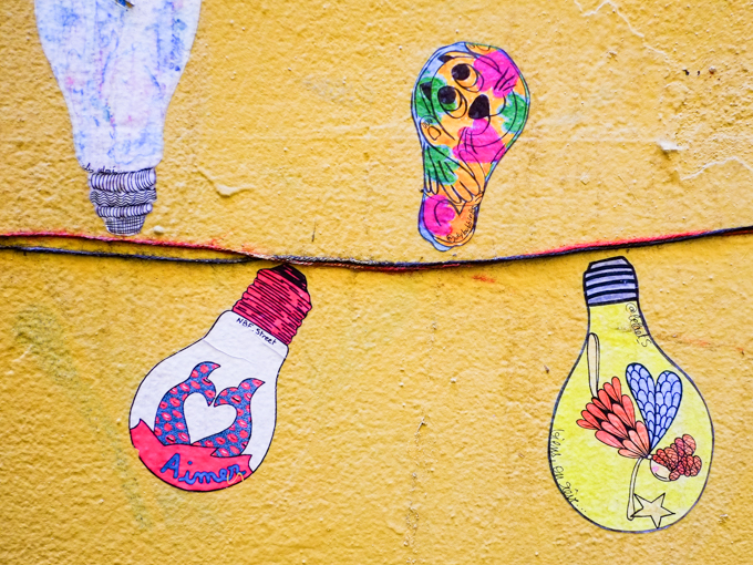 on a wall painted yellow, paste up graffiti of a string of light bulbs where each bulb is a little artwork by a different artist, 3 bulbs