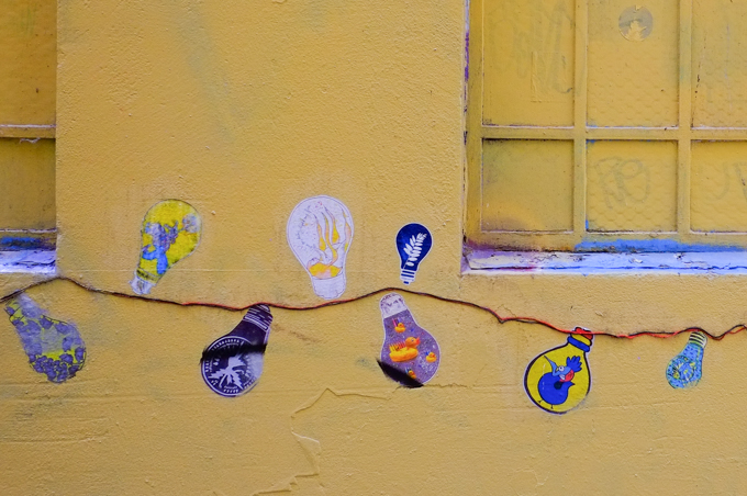 on a wall painted yellow, paste up graffiti of a string of light bulbs where each bulb is a little artwork by a different artist