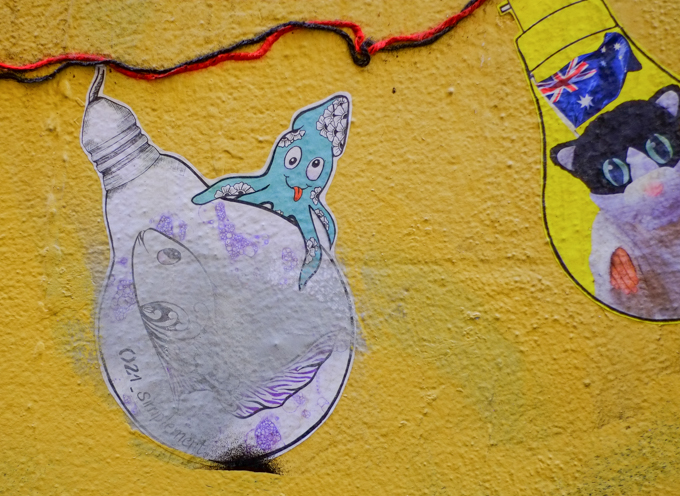paper pasteup on yellow wall, shape of a light bulb, a little blue octopus with its tongue sticking out, sits on top of one of the bulbs that contains a small fish. the other light bulb has a cat head, rest of bulb is out of the picture