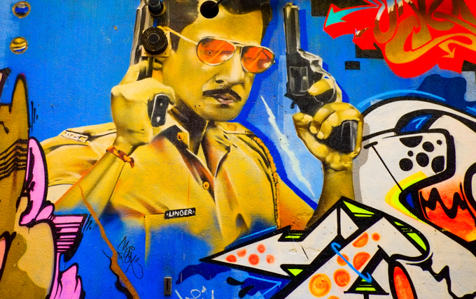 street art, part of a mural, man in orange sunglasses, a revolver in each hand, yellow jacket with name Unger written on it.