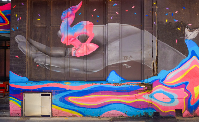 mural by vexta and lisa king, woman lying down on a rainbow of bright colours, on her back, eyes closed, hand outstretched with object in hand, in two sections, woman is in shades of grey